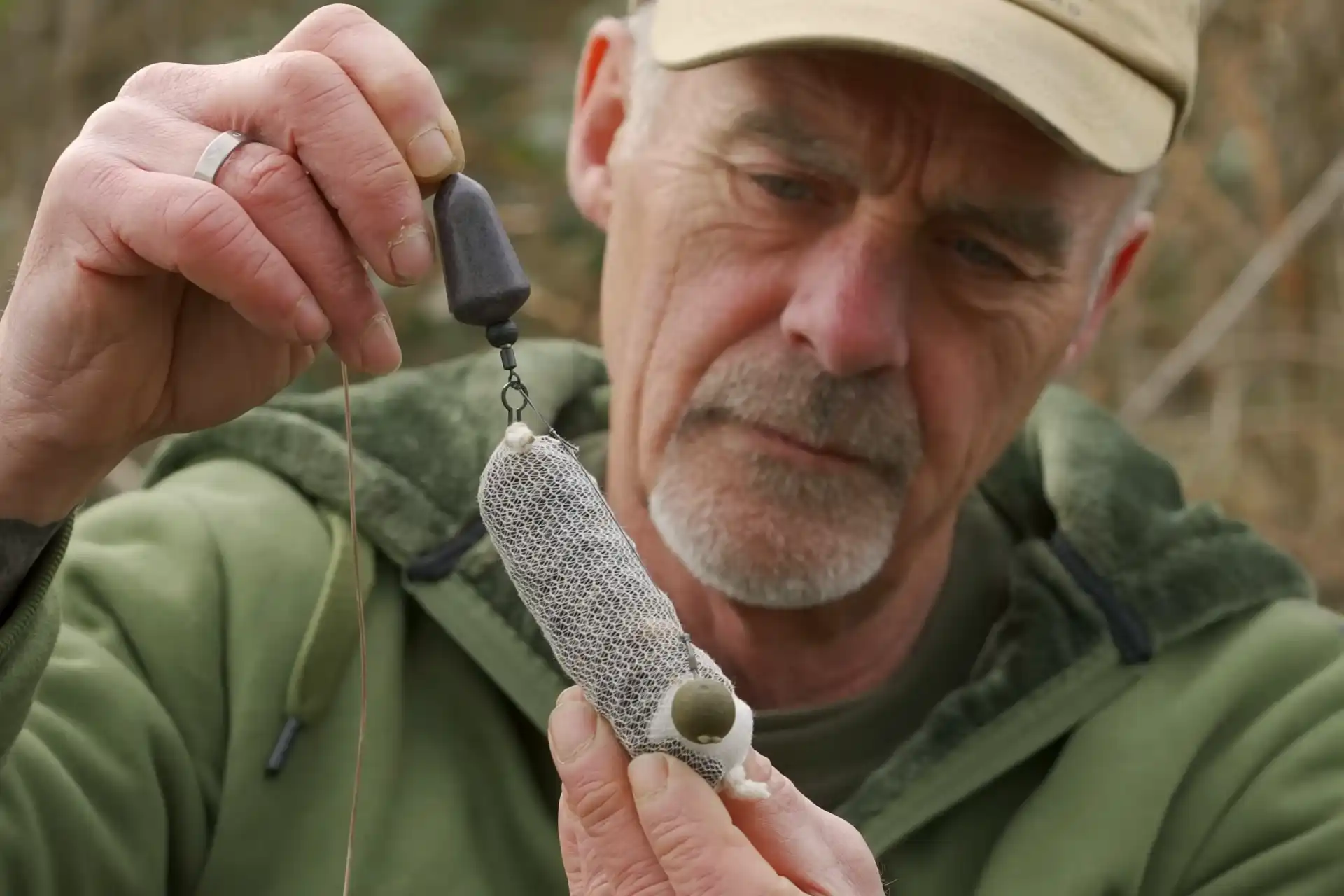 A person in outdoor clothing holds and examines a fishing rig, featuring a mesh bait bag attached to a fishing line with a weight.