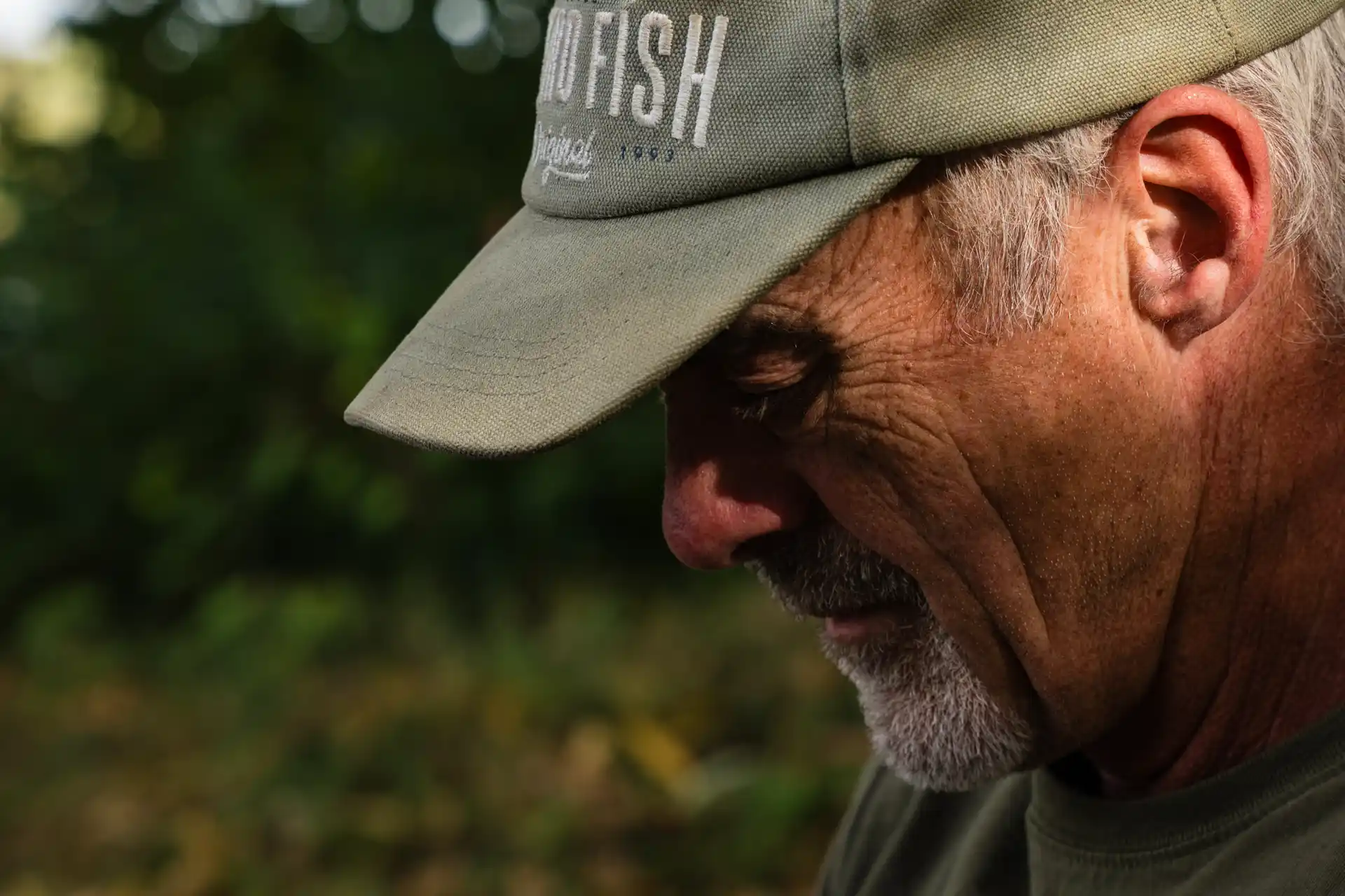 Close-up of an older man with a grey beard wearing a khaki cap, looking down thoughtfully. The background is blurred with greenery, and sunlight gently highlights his face and wrinkles.