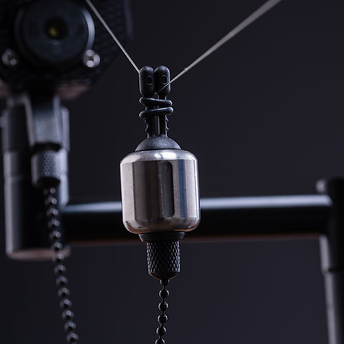 Close-up of Stainless Bug Bobbins, a metallic fishing bite indicator attached to a fishing line, with blurred fishing tackle in the background.