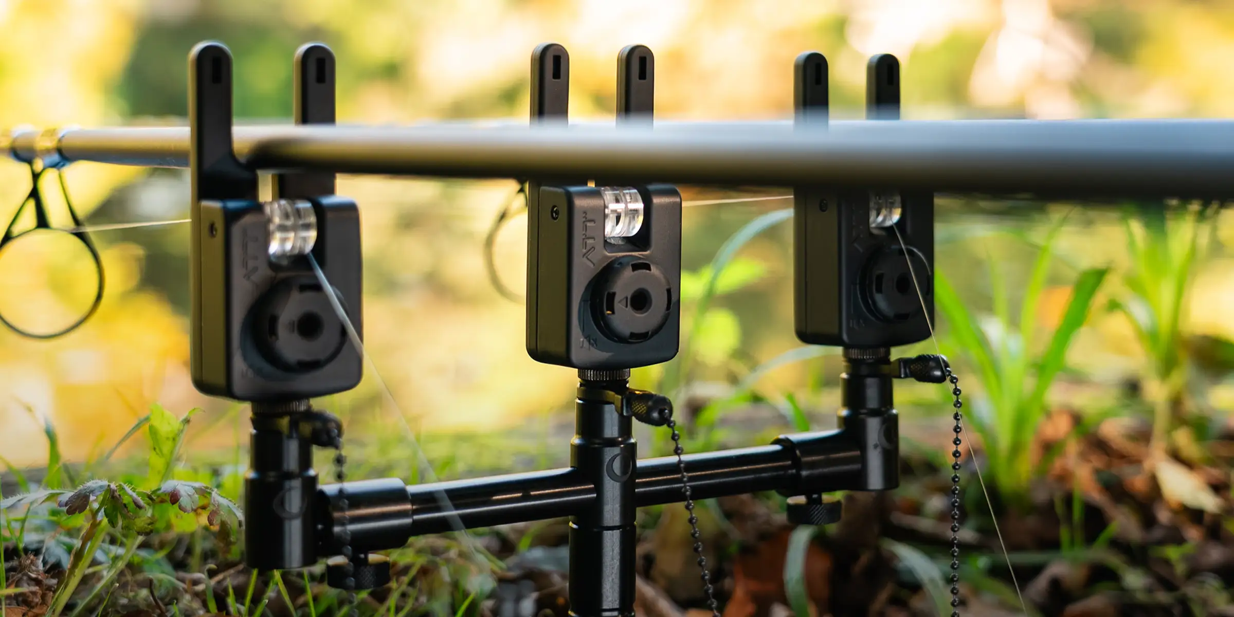Three black electronic bite alarms are mounted on a rod pod, set up outdoors on grass, with fishing lines threaded through each device and a blurred green background.