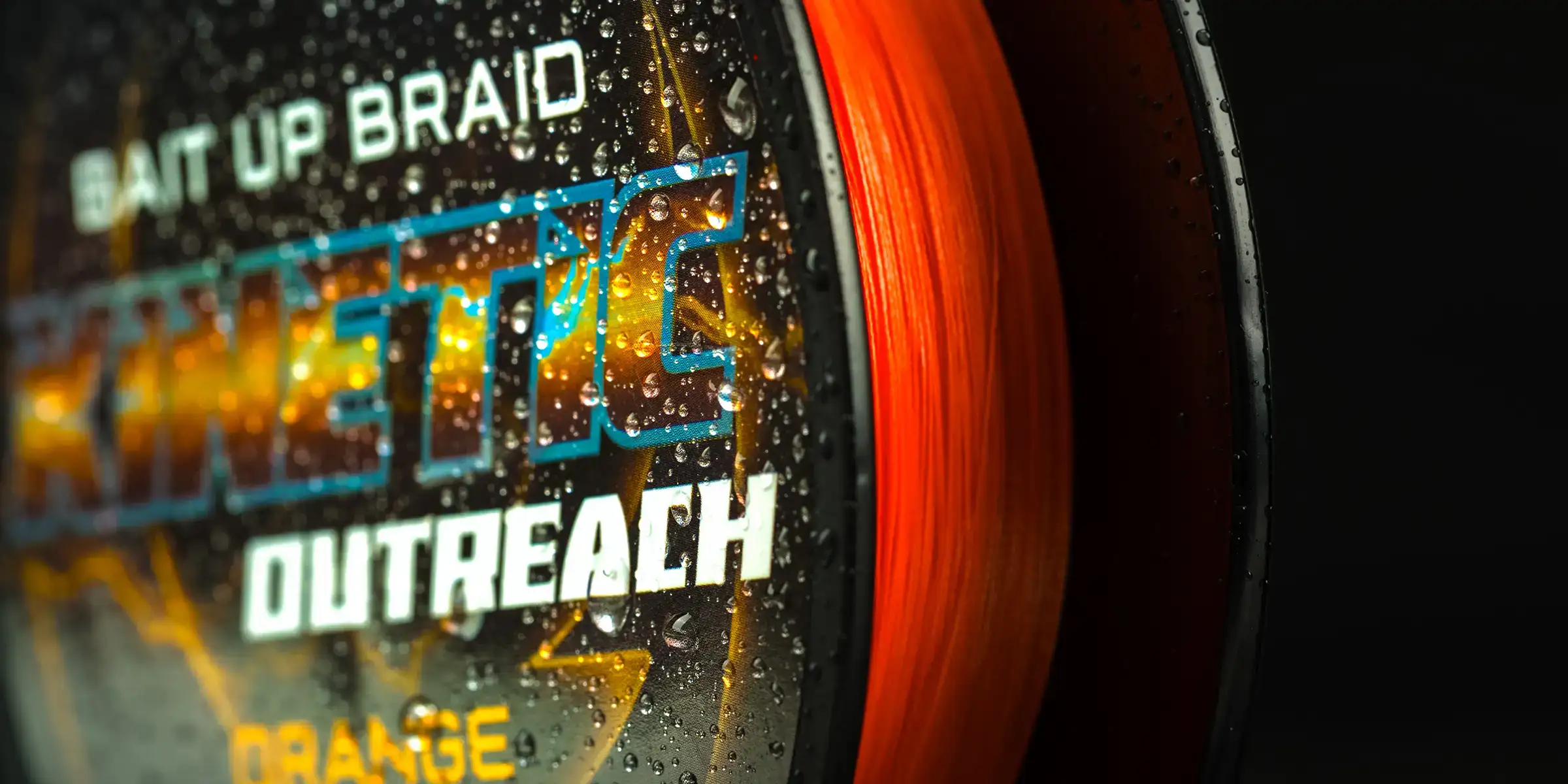 A close-up of a spool of orange fishing line labelled Kinetic Outreach Bait Up Braid, with water droplets on the packaging against a dark background.