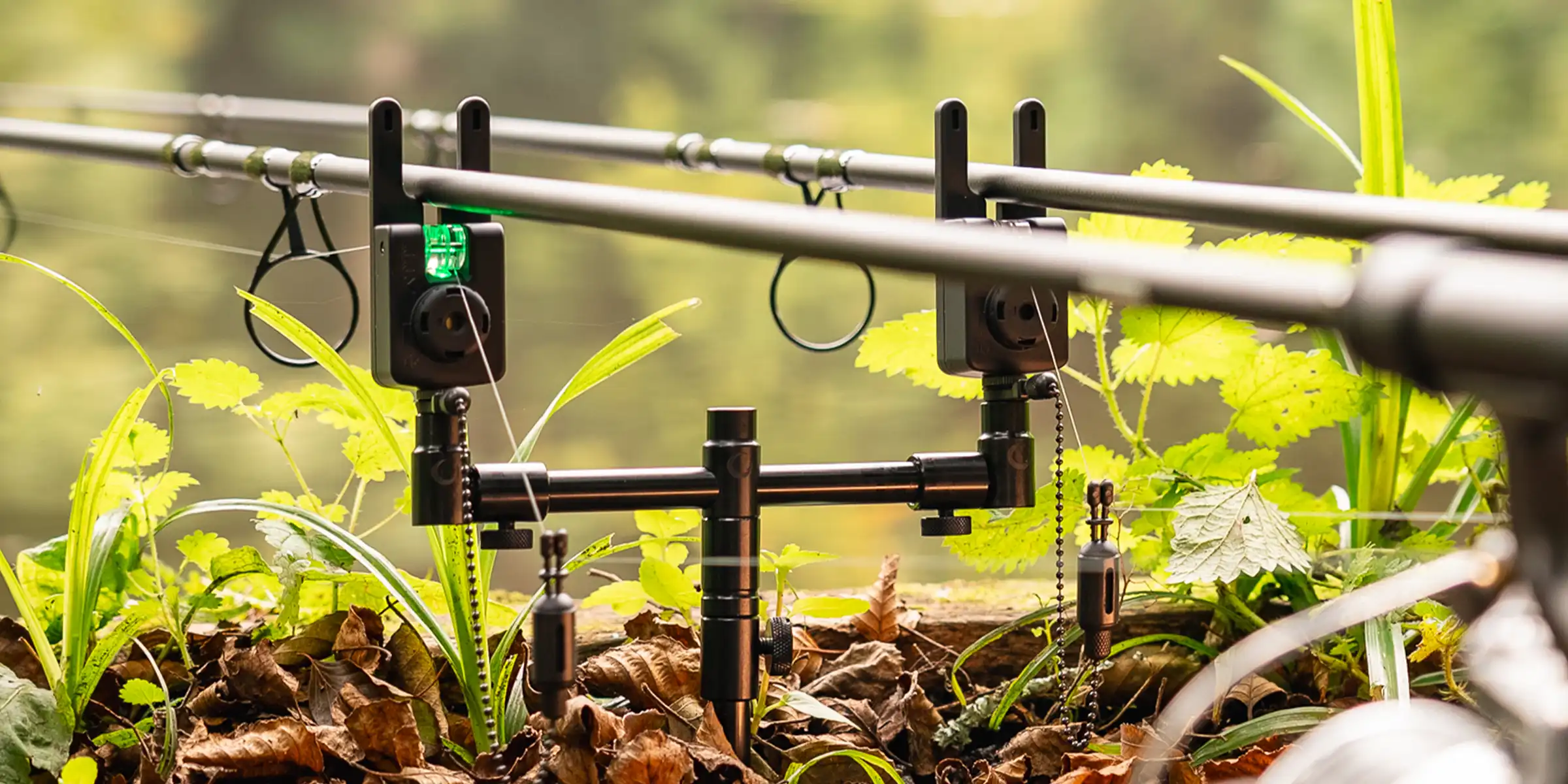Two fishing rods set up side by side with electronic bite alarms, positioned over leafy ground and surrounded by green plants, ready for angling in a natural outdoor setting.