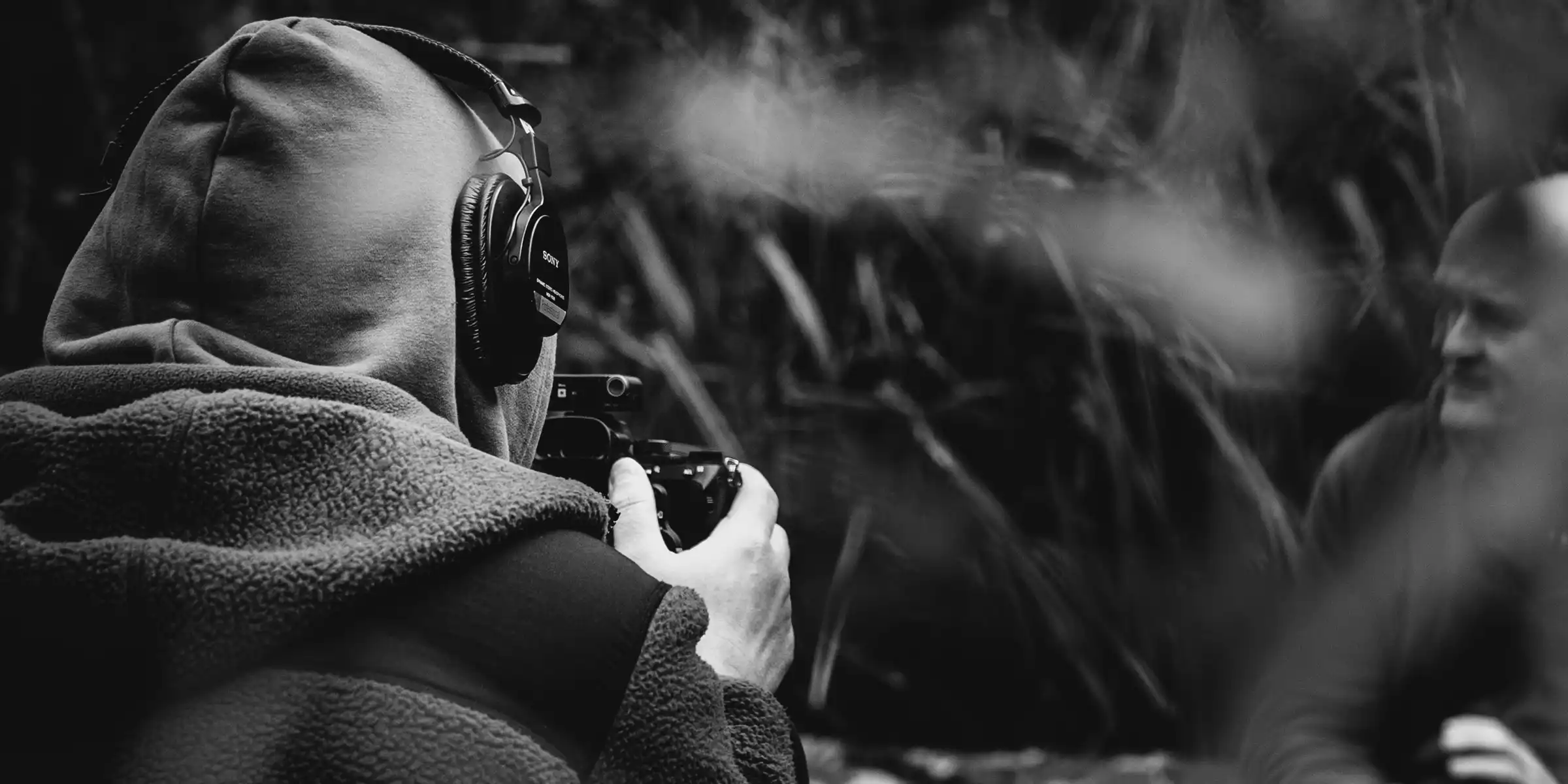 A person in a hoodie and headphones holds a camera, filming or photographing another person sitting outdoors. The image is in black and white, with a blurred natural background.