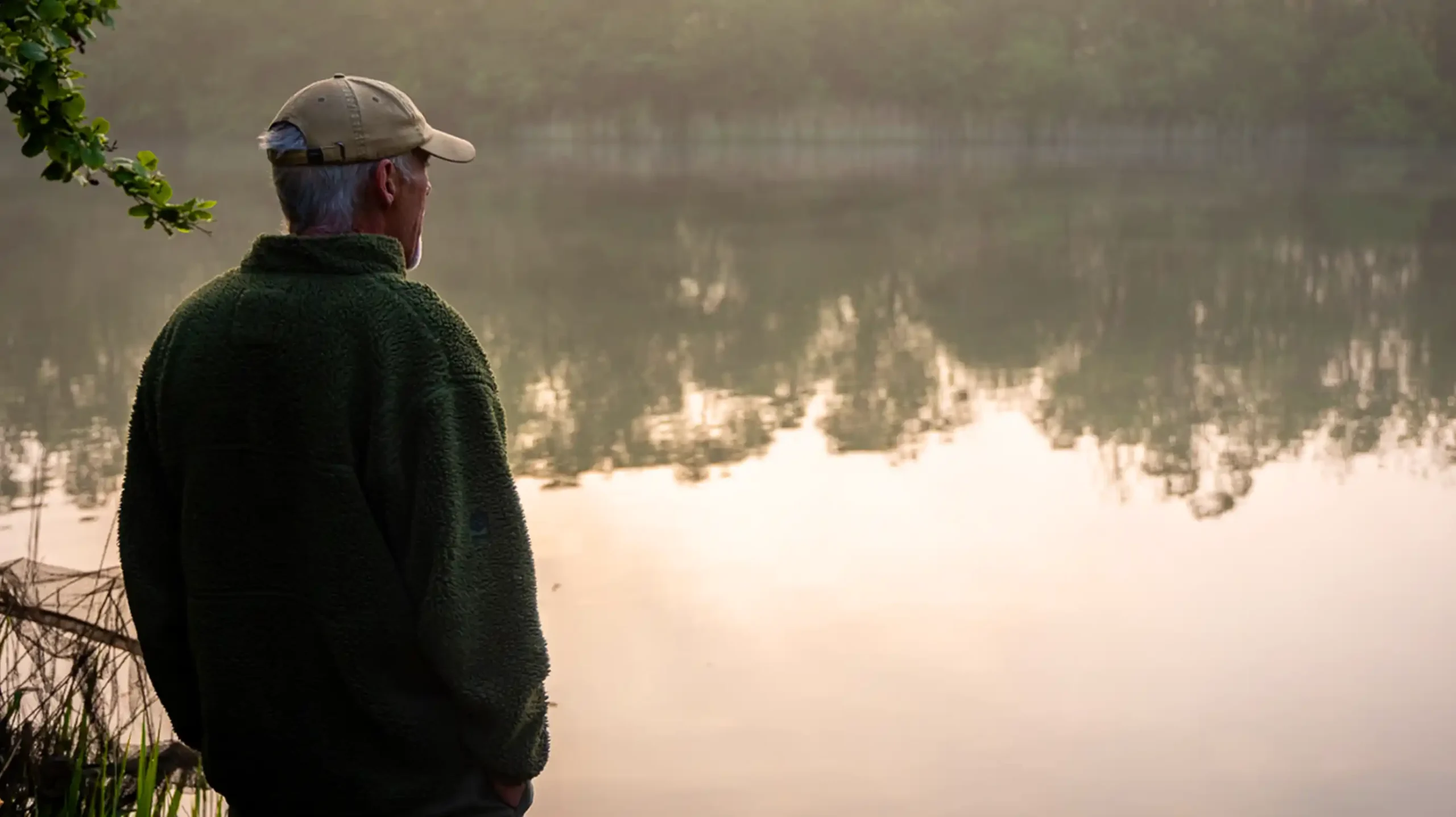 An older man wearing a cap and a green jacket stands by a calm lake, gazing at the water with trees and mist reflected on the surface in the early morning light.