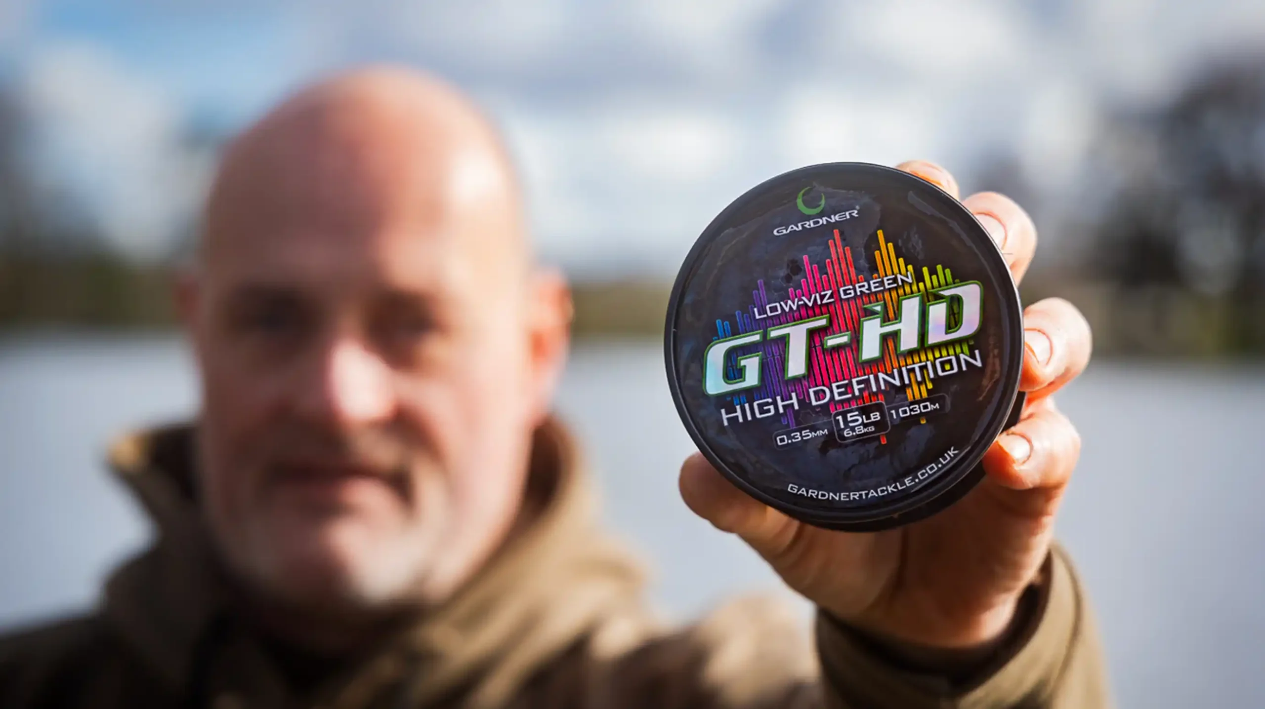 A person outdoors holds a round container labelled GT-HD High Definition Lowviz Green fishing line, with the label in sharp focus and the person's face blurred in the background.