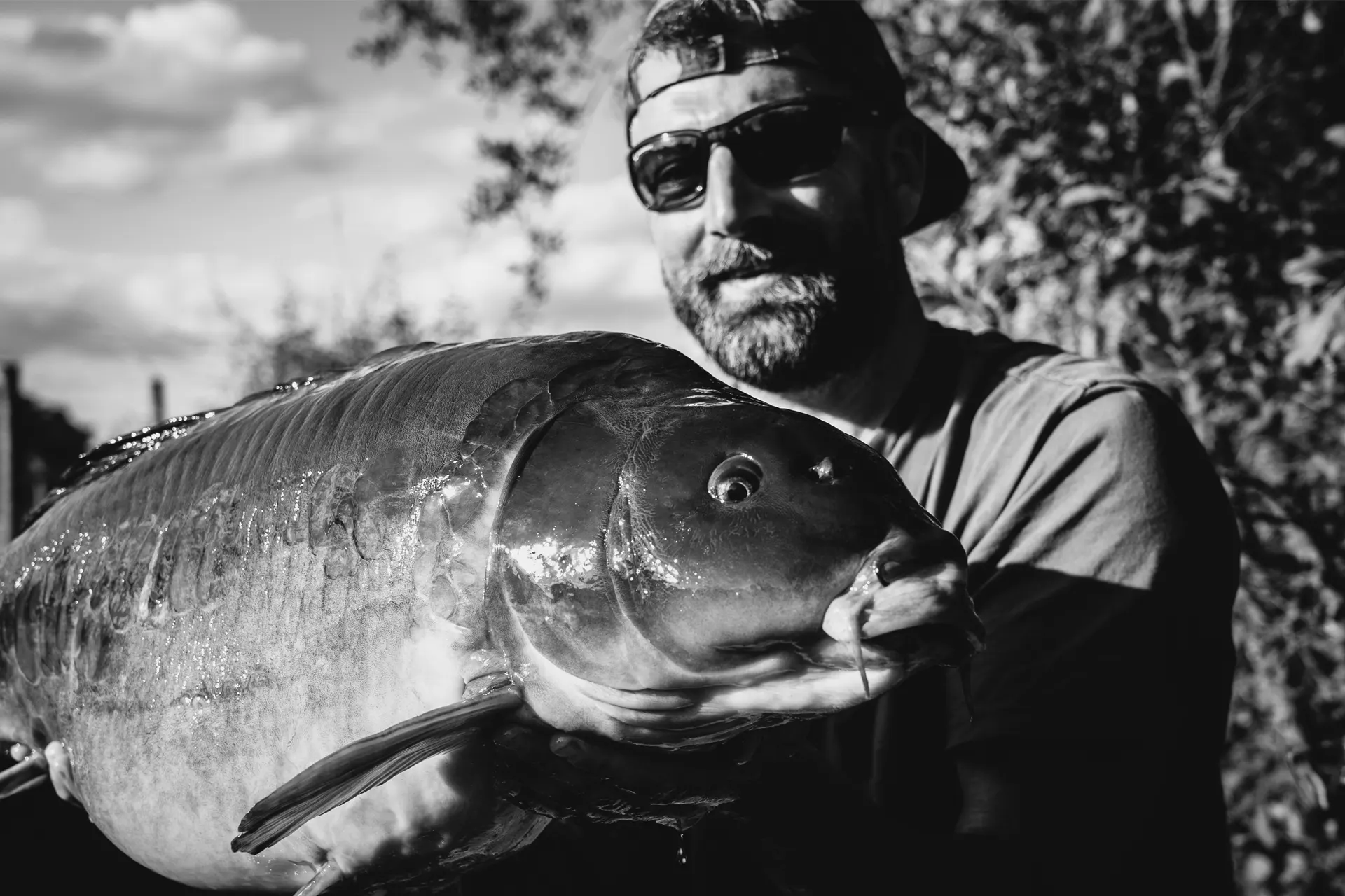 A man wearing sunglasses and a backwards cap smiles whilst holding a large fish outdoors. The image is in black and white, with trees and a cloudy sky in the background.