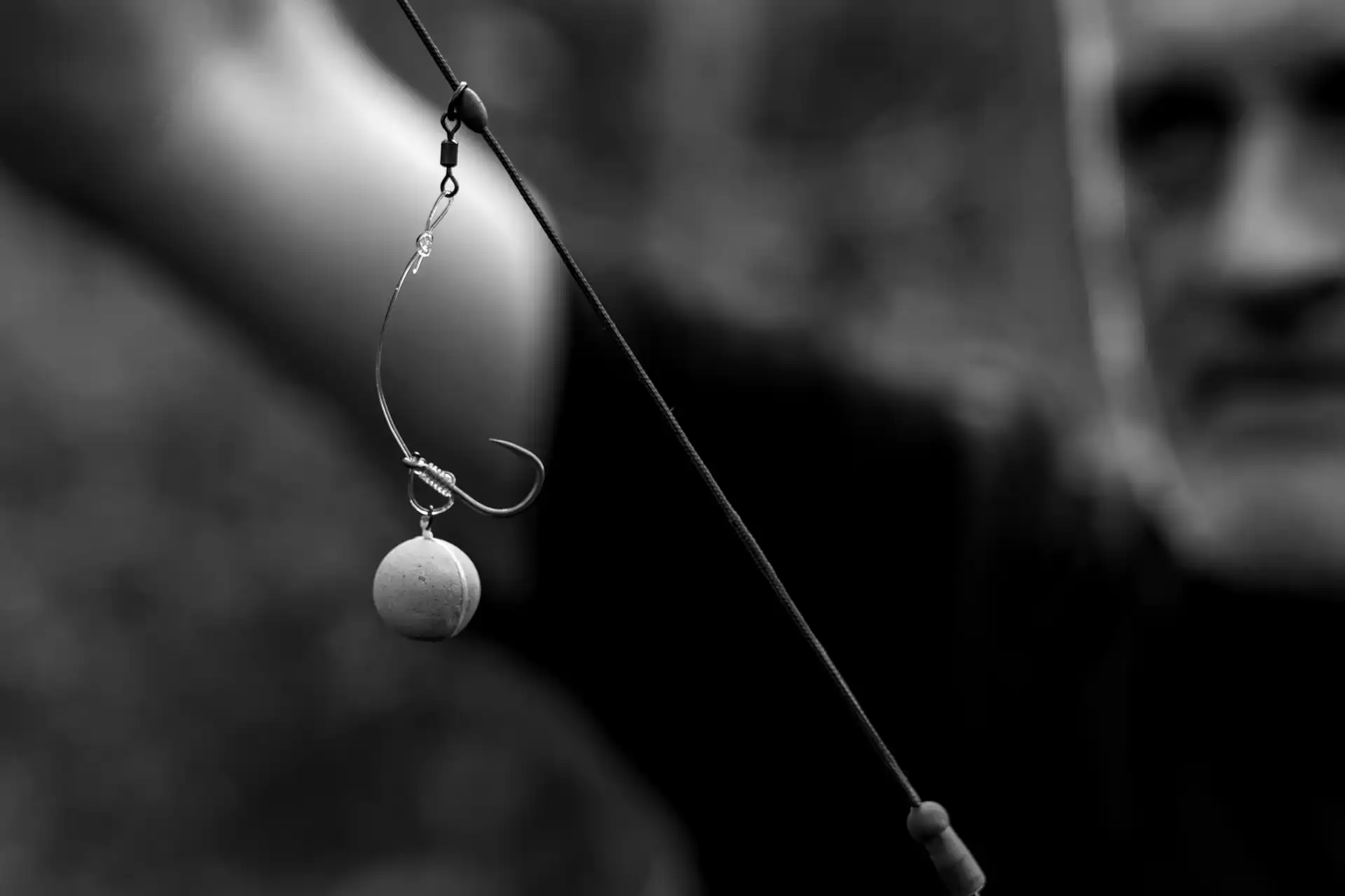 A close-up black-and-white photo of a fishing hook with bait attached, held by a person whose face is blurred in the background.