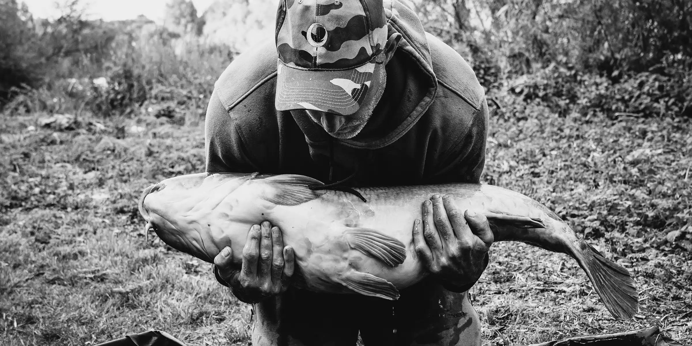 A person wearing a camouflage cap and hoodie is bending forward, holding a large fish with both hands outdoors. The scene is in black and white, with blurred trees and grass in the background.